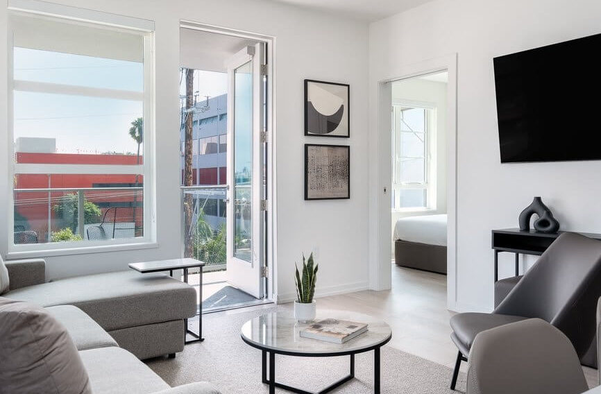 Modern living room with neutral-toned furniture, a round coffee table, wall art, and a large TV. Glass door opens to a balcony with city view. A bedroom is visible through an open doorway.