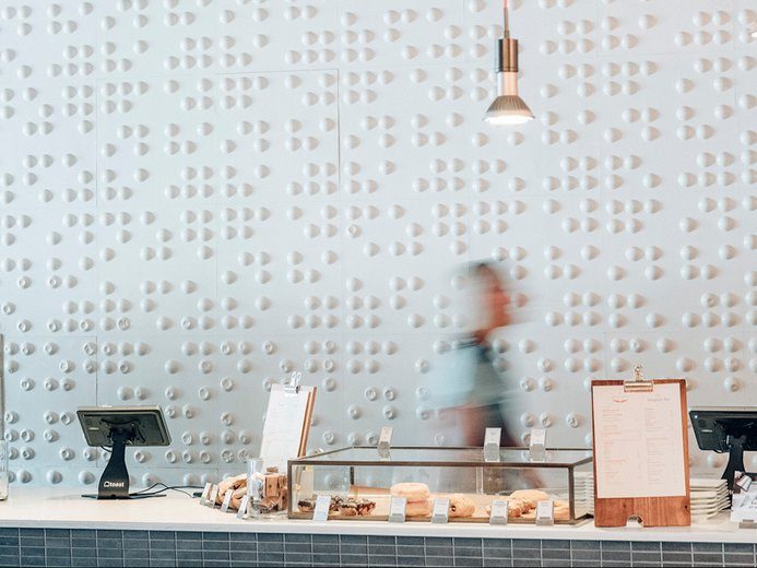 A blurred person walks behind a counter with pastries, two registers, and menus in a modern cafe with a textured white wall and a hanging light above.