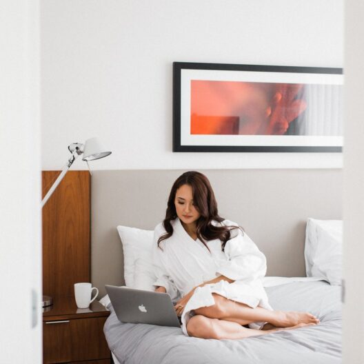 A woman in a white robe sits on a neatly made bed using a laptop. A mug and lamp are on the bedside table, and a modern artwork hangs on the white wall above her.