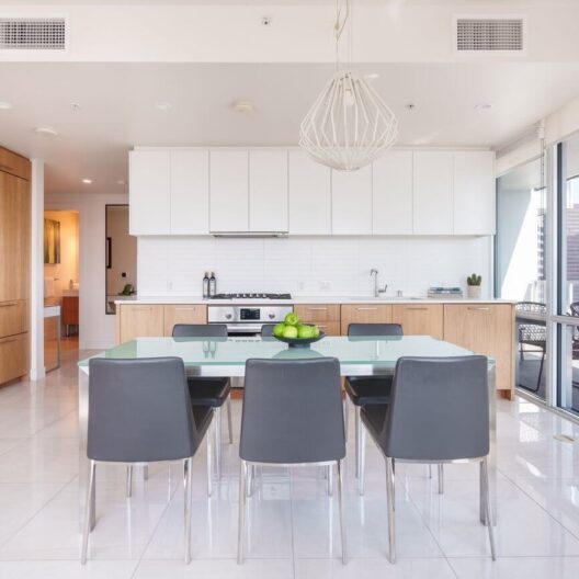 Modern kitchen with white cabinets, light wood lower cabinets, a glass dining table with six gray chairs, pendant light above, and a bowl of green apples on the table. Large windows bring in natural light.