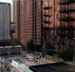 A rooftop pool with lounge chairs and cabanas is surrounded by tall brick and glass apartment buildings in a city setting, lit by soft evening sunlight.