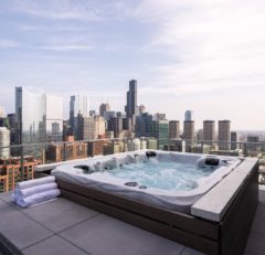 A rooftop hot tub with bubbling water sits beside neatly rolled white towels, overlooking a city skyline with tall modern buildings under a partly cloudy sky.