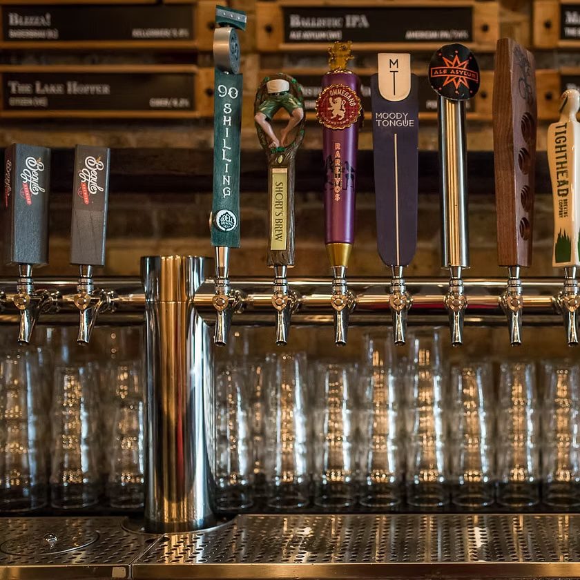 A row of beer taps with unique handles, set above a counter and in front of stacked clean glassware, with a wooden menu board displaying beer names in the background.