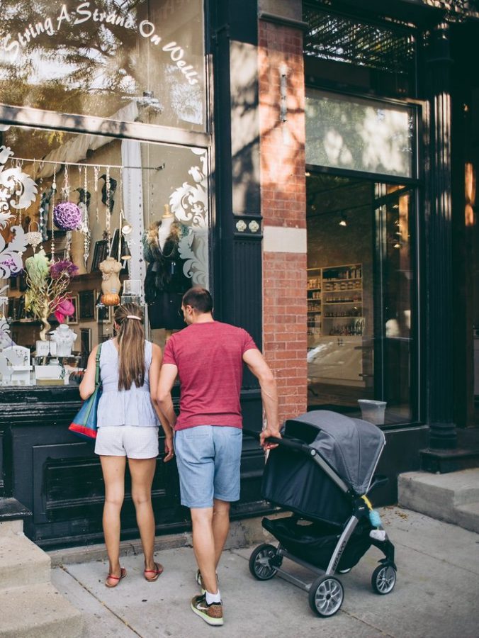 A couple stands outside a shop window on a city sidewalk, holding hands. The man pushes a baby stroller while both look at items displayed in the store.
