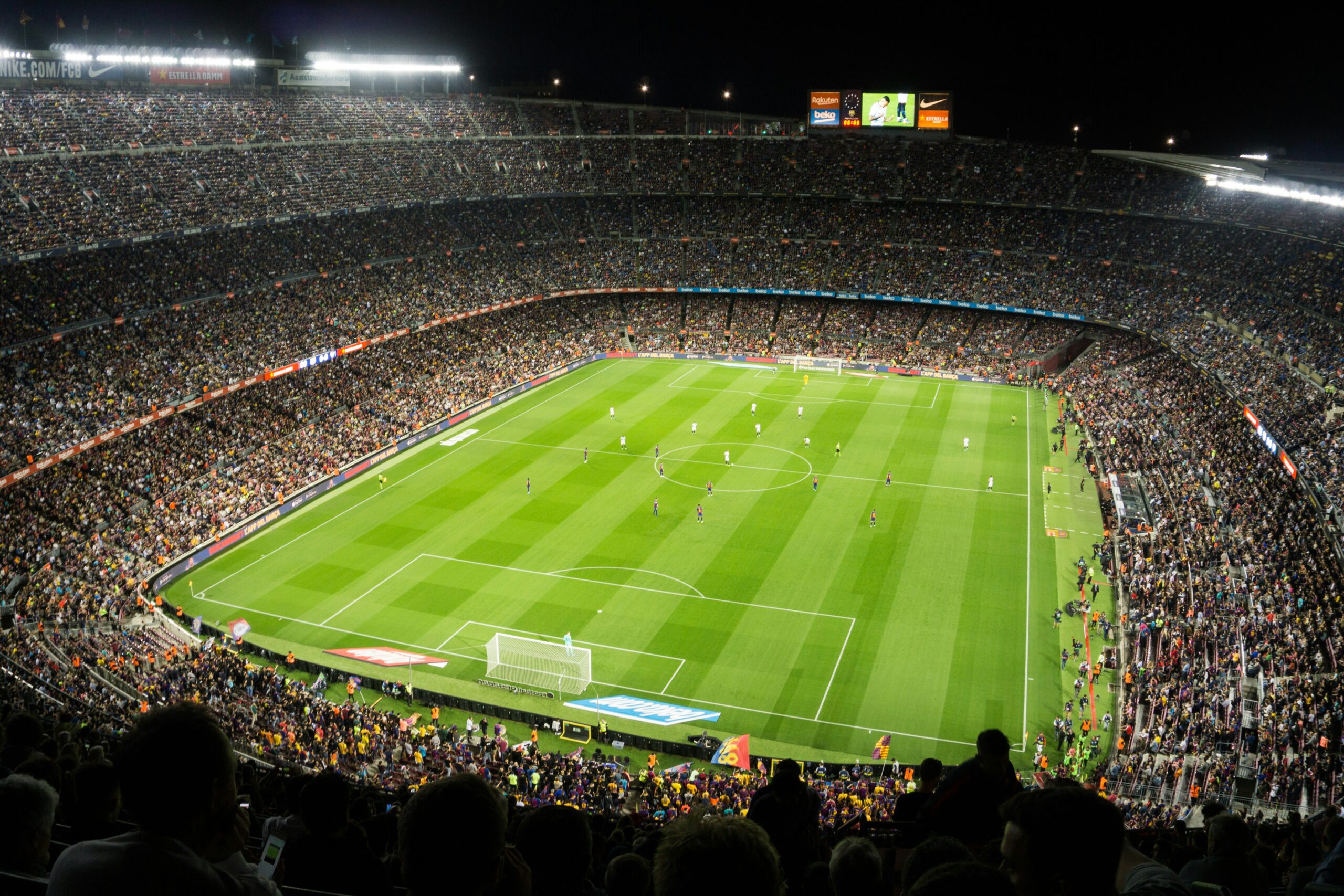 A packed football stadium at night, filled with spectators watching a match on a brightly lit green field; players are spread across the pitch, and large advertising boards line the sidelines.