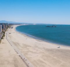 Aerial view of a wide sandy beach with a walking path, calm blue ocean, and distant city buildings under a clear blue sky. A few small boats are visible on the water.