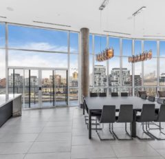 A modern dining area with a large black table, gray chairs, pendant lights, floor-to-ceiling windows, and a city skyline view in the background. Light gray tile flooring and a tall concrete pillar are visible.