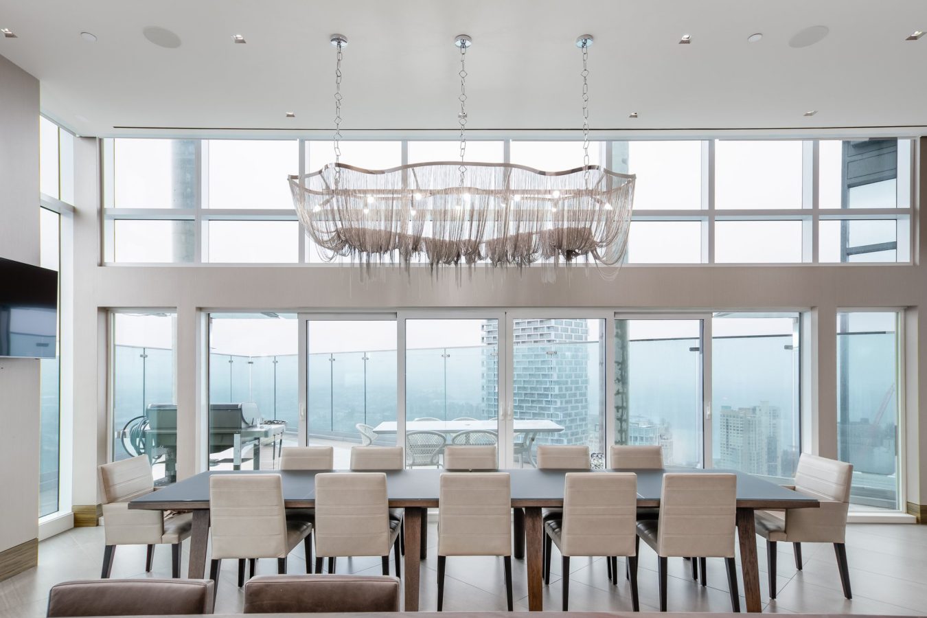 A modern dining room with a long table and ten beige chairs, large floor-to-ceiling windows revealing a city view, and a contemporary chandelier hanging above the table.