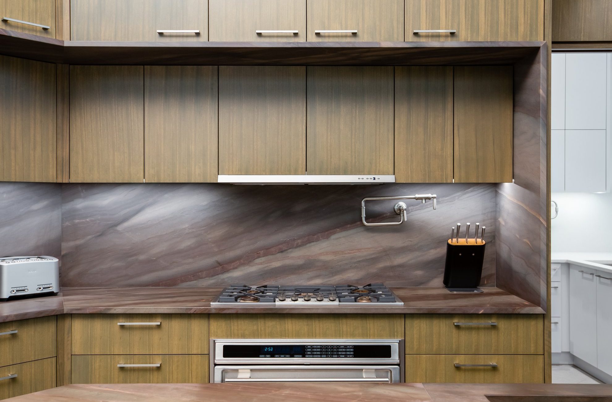 Modern kitchen with wood cabinets, marble backsplash and countertop, stainless steel gas stove, built-in oven, toaster on the left, knife block on the right, and a wall-mounted pot filler above the stove.