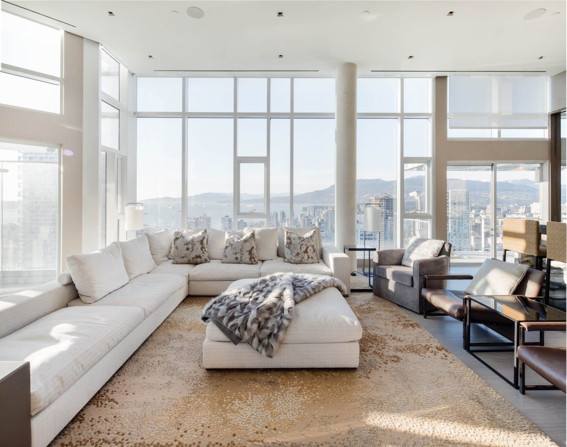 Bright, modern living room with large windows, white sectional sofa, throw blanket, patterned rug, and cityscape mountain view in the background. Neutral tones, abundant natural light, and contemporary furnishings.