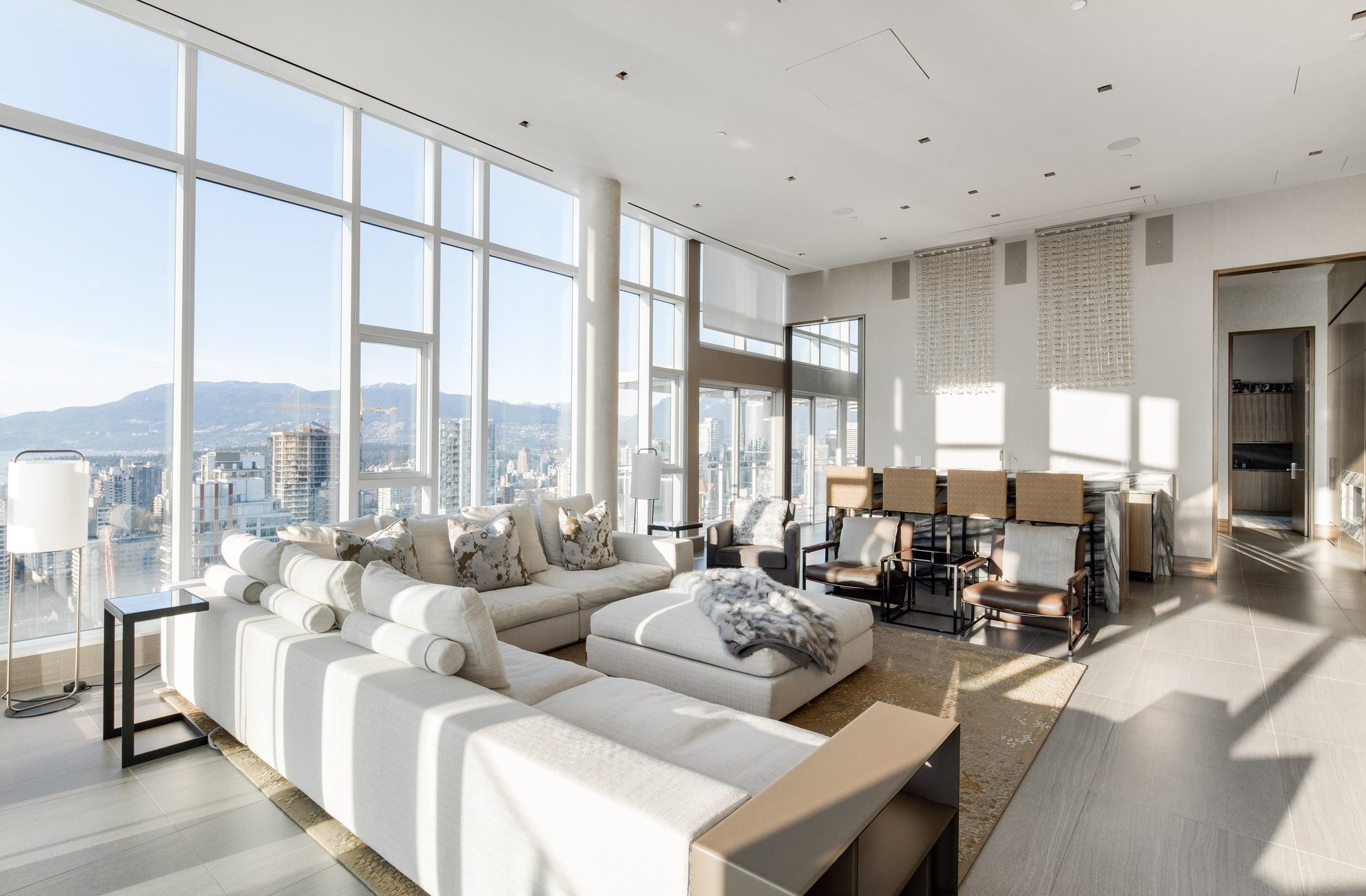 A spacious, modern living and dining area with floor-to-ceiling windows offering city and mountain views, featuring light-colored sofas, a large dining table, and minimalist decor bathed in natural sunlight.