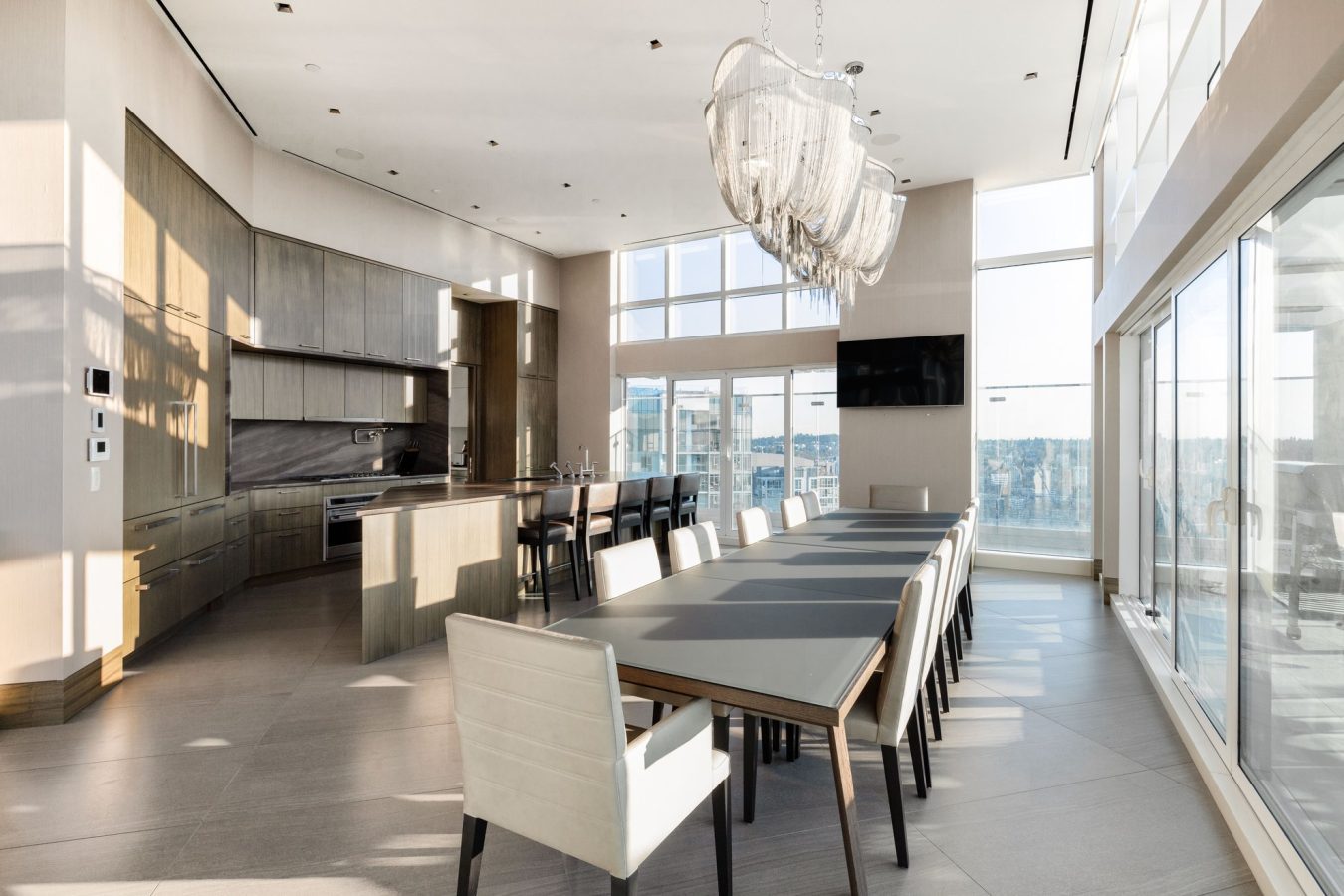 A modern dining room with a long table, white chairs, large windows with city views, a contemporary chandelier, and an open kitchen area with sleek cabinetry in the background.