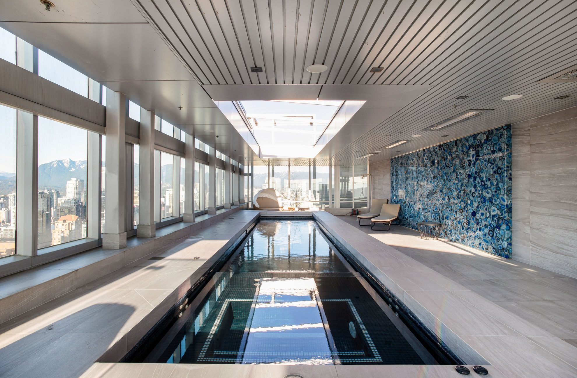A modern indoor lap pool with floor-to-ceiling windows, mountain and city views, a blue mosaic accent wall, and lounge chairs, all under a slatted ceiling with a large skylight.