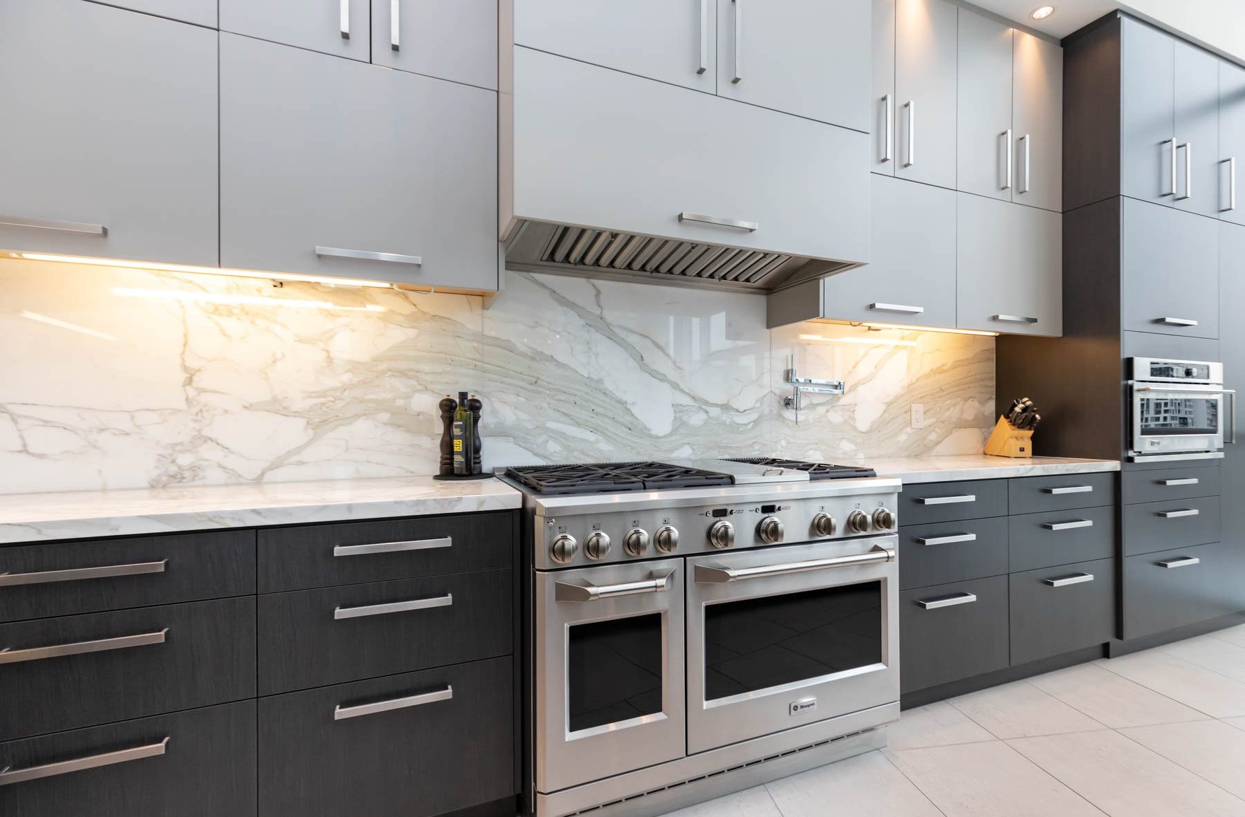 Modern kitchen with gray cabinets, marble backsplash, built-in oven, stainless steel gas stove, under-cabinet lighting, a knife block, pepper grinder, and utensil holder on white countertops.