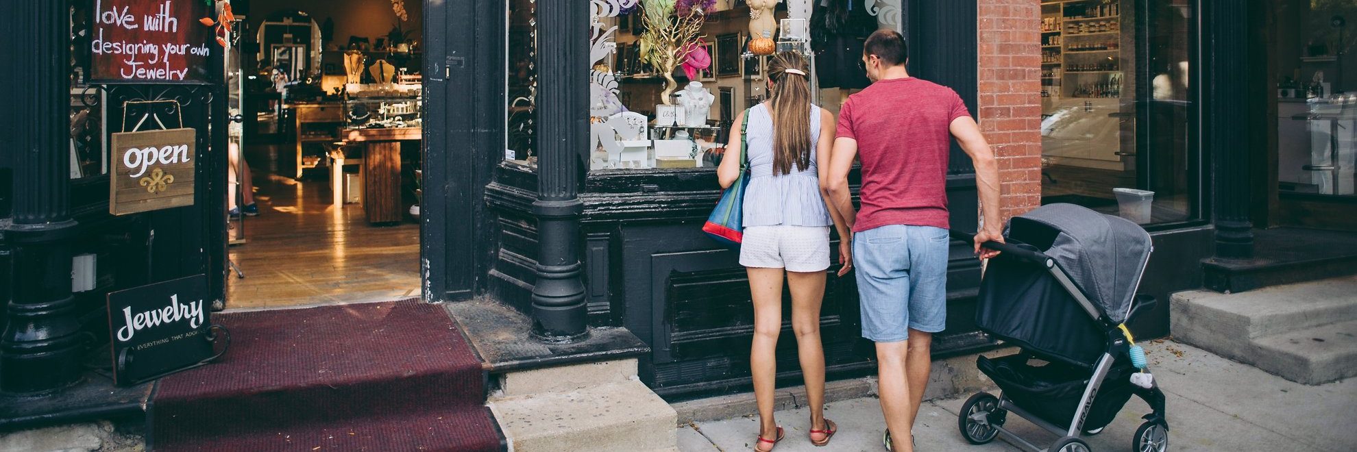 A couple, seen from behind, stands outside a jewelry store window with a stroller, looking at the displays. The man wears a red shirt and shorts; the woman wears a white outfit and hat. The store has an “open” sign displayed.