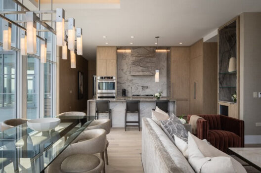 Modern open-concept kitchen and living area with light wood cabinets, marble backsplash, a central island with stools, a glass dining table, upholstered chairs, and contemporary pendant lighting.
