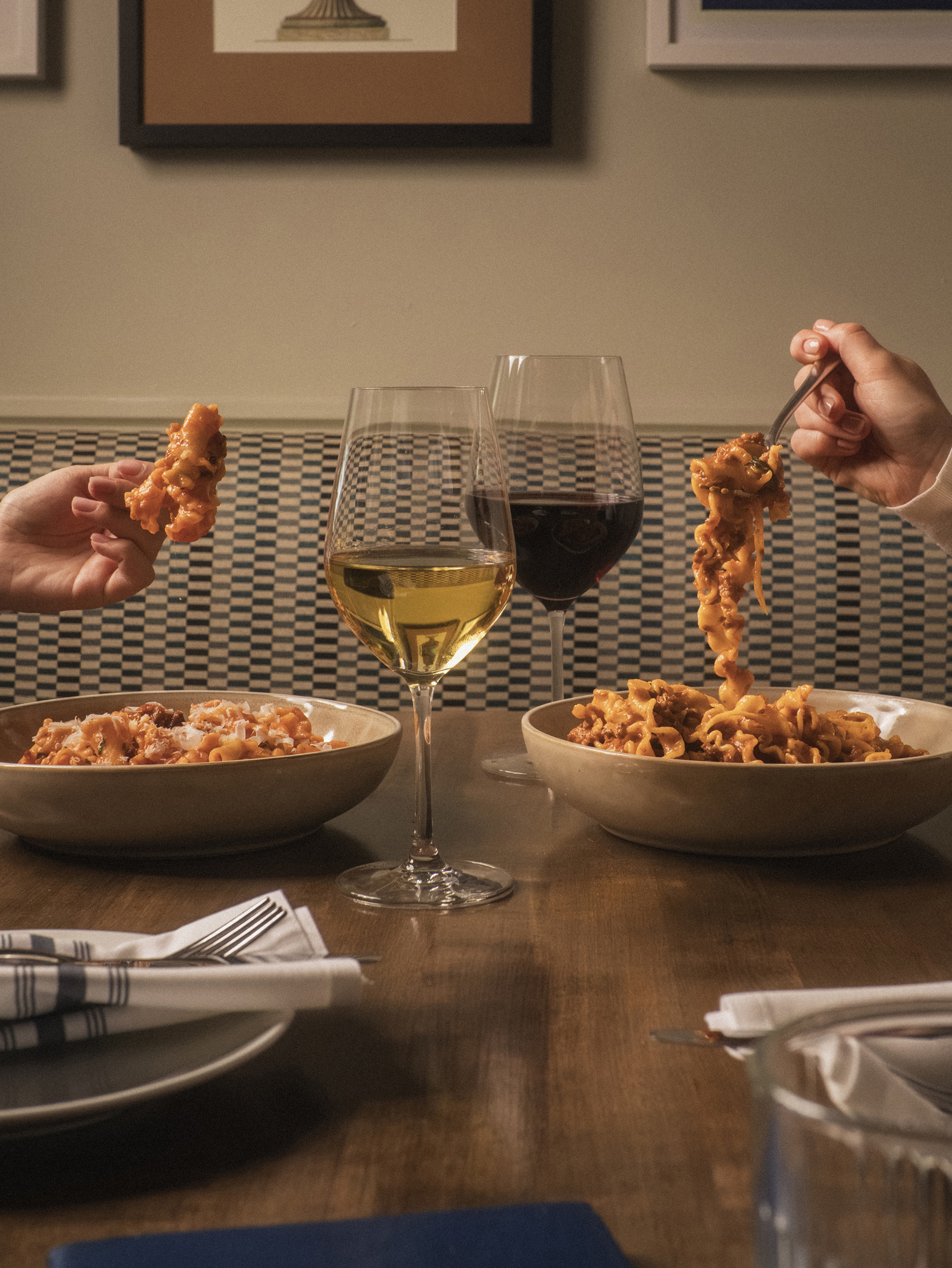 Two people hold forkfuls of pasta above bowls on a wooden table set with napkins, a glass of white wine, and a glass of red wine, with framed art on the wall in the background.