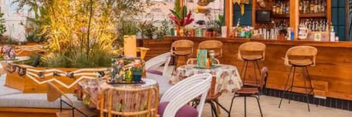 Outdoor restaurant patio with round tables, floral tablecloths, wicker chairs, and a wooden bar lined with stools. Green plants and warm lighting create a cozy, inviting atmosphere.