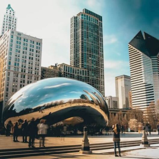 People stand near the reflective Cloud Gate sculpture, known as The Bean, in Chicagos Millennium Park, surrounded by tall skyscrapers under a partly cloudy sky.