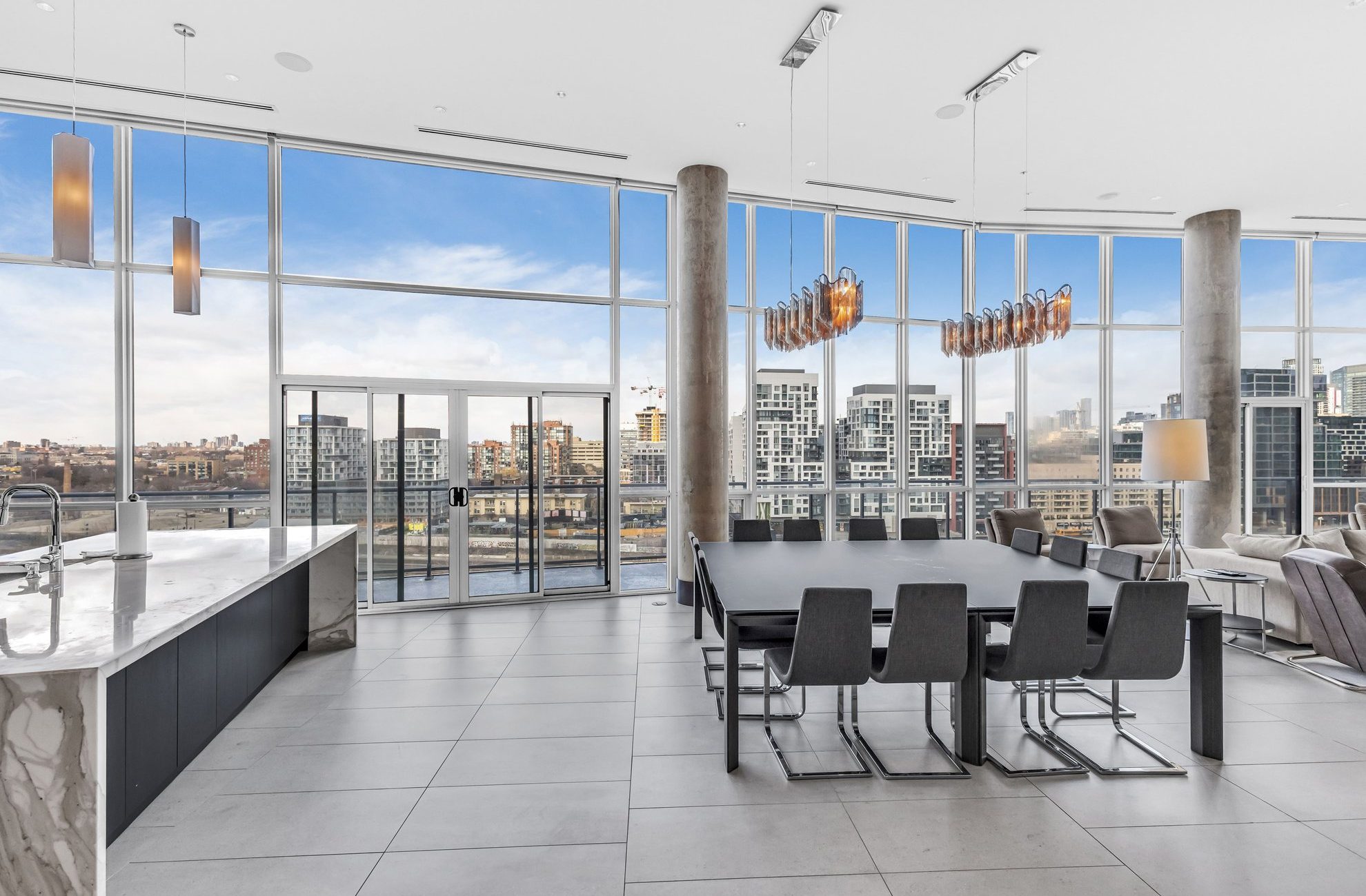 Modern open-concept kitchen and dining area with large floor-to-ceiling windows, city skyline views, marble countertops, and contemporary lighting fixtures. Black dining chairs surround a long table.