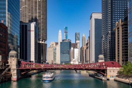 A tour boat passes under a red steel bridge on the Chicago River, surrounded by tall modern skyscrapers and historic buildings on a clear, sunny day.