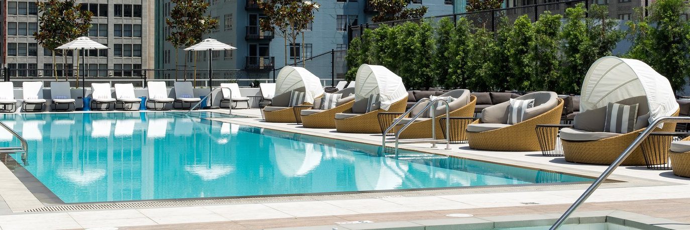 A rooftop swimming pool surrounded by lounge chairs and round wicker daybeds with umbrellas, set against a backdrop of tall buildings and greenery.