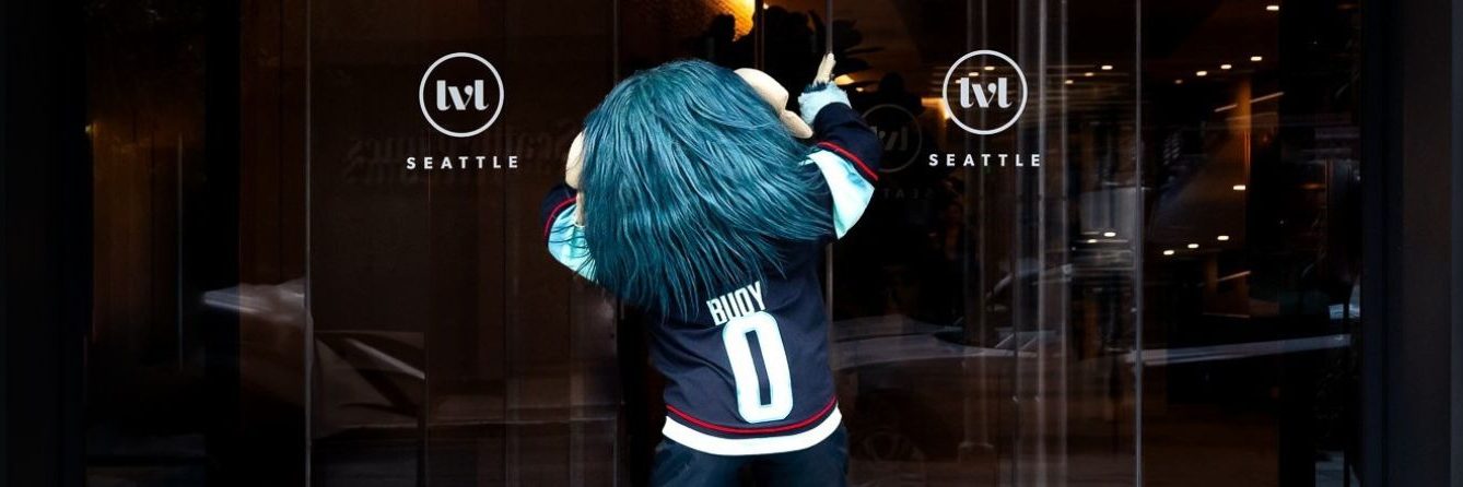 A mascot with long blue hair and a jersey labeled BUOY 0 faces glass doors at a building entrance with ivt SEATTLE logos on each door.