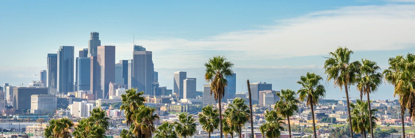 Los Angeles city skyline with tall modern buildings and palm trees in the foreground under a blue sky with scattered clouds.