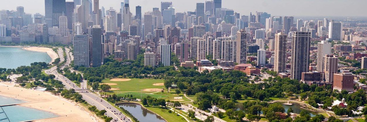 Aerial view of downtown Chicago with Lake Michigan on the left, green parks and baseball fields in the center, and tall skyscrapers filling the city skyline under a hazy sky.