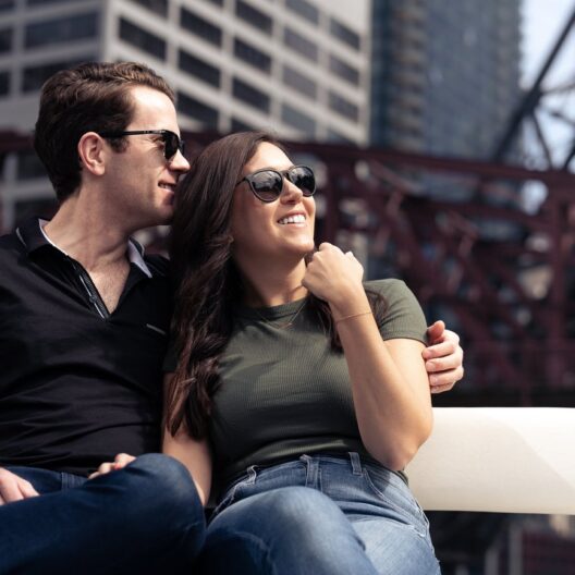 A smiling couple wearing sunglasses sits close together on a bench outdoors, with city buildings and a red steel bridge blurred in the background.