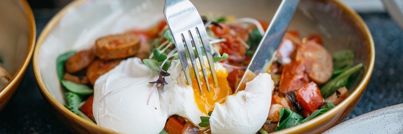 A fork and knife cut into a poached egg on a salad with sliced sausage, greens, and diced red vegetables in a brown-rimmed bowl. The egg yolk is oozing onto the other ingredients.