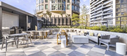 Modern outdoor patio with checkered flooring, various tables and chairs, cushioned seating, and built-in grills, surrounded by planters and tall apartment buildings in the background on a sunny day.