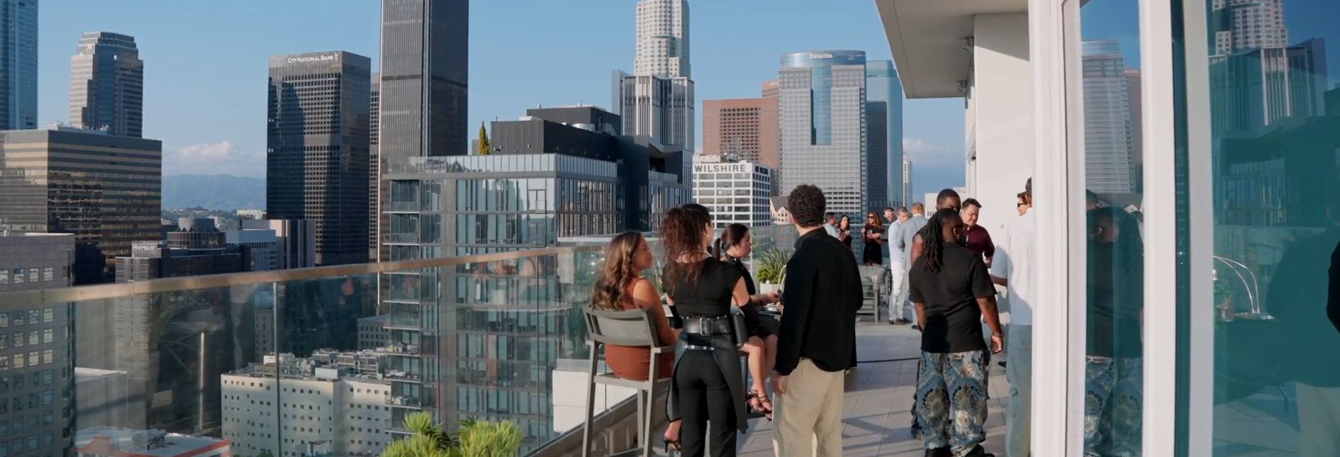 People gather and socialize on a high-rise balcony overlooking a city skyline with tall buildings, glass railings, and clear blue skies in the background.