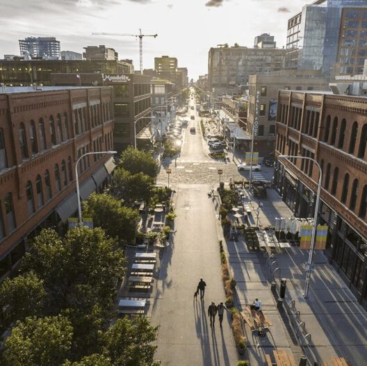 A wide, sunlit city street lined with red brick buildings, outdoor seating, trees, and people walking. Modern buildings and construction cranes are visible in the background under a partly cloudy sky.