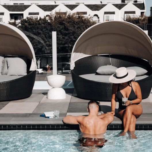 A man sits in a pool holding hands with a woman sitting poolside. The woman wears a hat and holds a drink. Behind them are round shaded lounge beds with cushions. A book and glass are on the edge of the pool.
