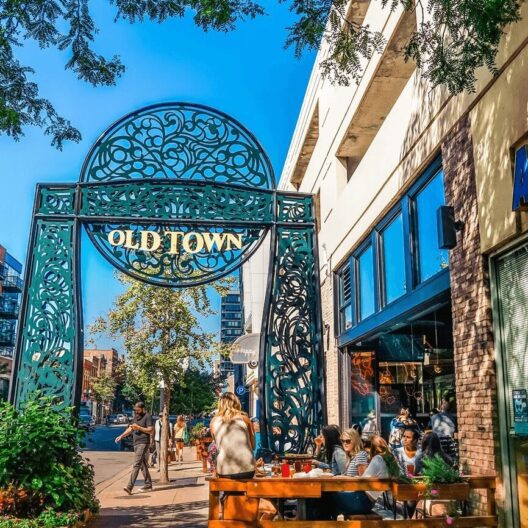 A group of people sit at an outdoor table near a restaurant under a large ornate metal “Old Town” sign on a sunny day, with trees, buildings, and pedestrians in the background.