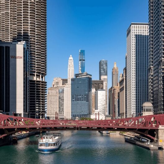 A boat travels along the Chicago River beneath a red bridge, surrounded by tall skyscrapers under a clear blue sky.