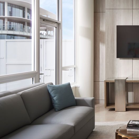 A modern living room with a gray sofa, a blue cushion, large windows, a wall-mounted TV, a wooden table, and light-colored walls and floors. Natural light fills the space.