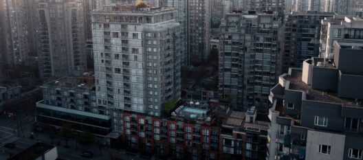 Aerial view of tall apartment buildings in a city, with a rooftop swimming pool visible in the center among the gray high-rises. Streets and parked cars are seen below, and the urban area appears densely populated.