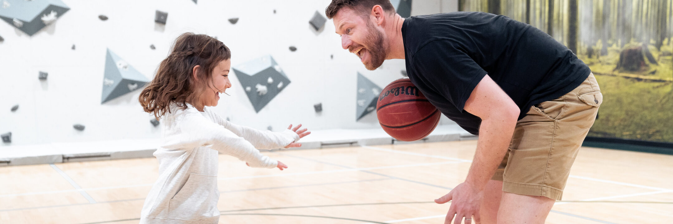 A young girl and a man play basketball indoors. The man, holding a basketball, leans forward and smiles at the girl, who reaches out toward him with excitement. The background features a climbing wall and forest mural.