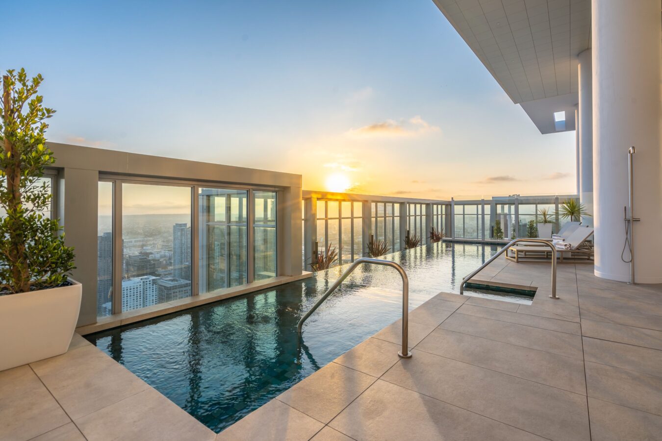 A rooftop infinity pool with glass railings overlooks a city skyline at sunset. Lounge chairs and potted plants line the deck, creating a luxurious and relaxing atmosphere.