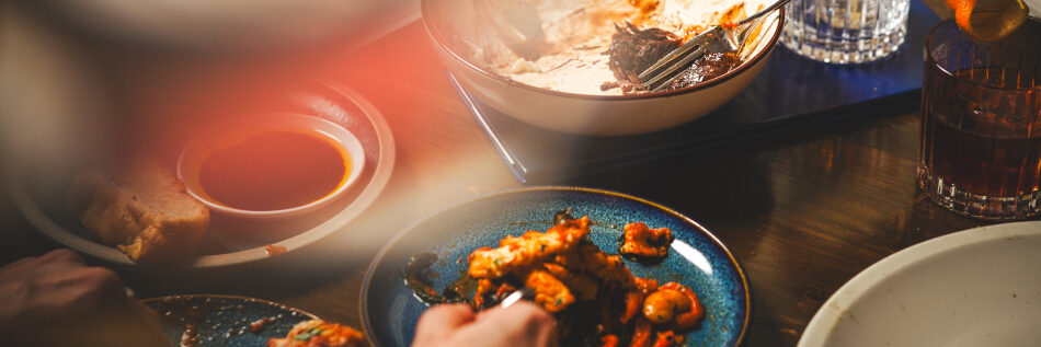 A close-up of a dinner table with plates of food, a bread roll, dipping sauce, drinks in glasses, and someones hand reaching for food. The scene is warmly lit with a cozy, casual atmosphere.
