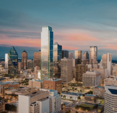 Aerial view of a city skyline at sunset, featuring tall modern skyscrapers, illuminated buildings, and a mix of architectural styles under a partly cloudy sky.