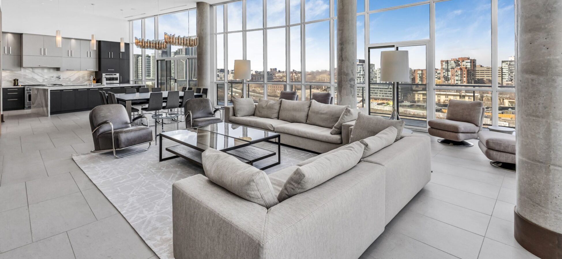 Spacious modern living room with large beige sectional sofa, glass coffee table, and floor-to-ceiling windows offering city skyline views. Open-concept layout includes a sleek kitchen and dining area with contemporary furnishings.