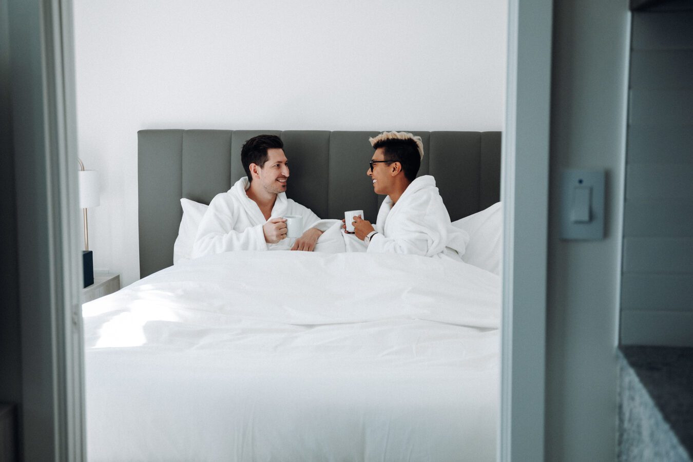 Two people in white bathrobes sit up in bed, smiling and holding mugs, in a bright, modern bedroom with white bedding and minimalist decor.