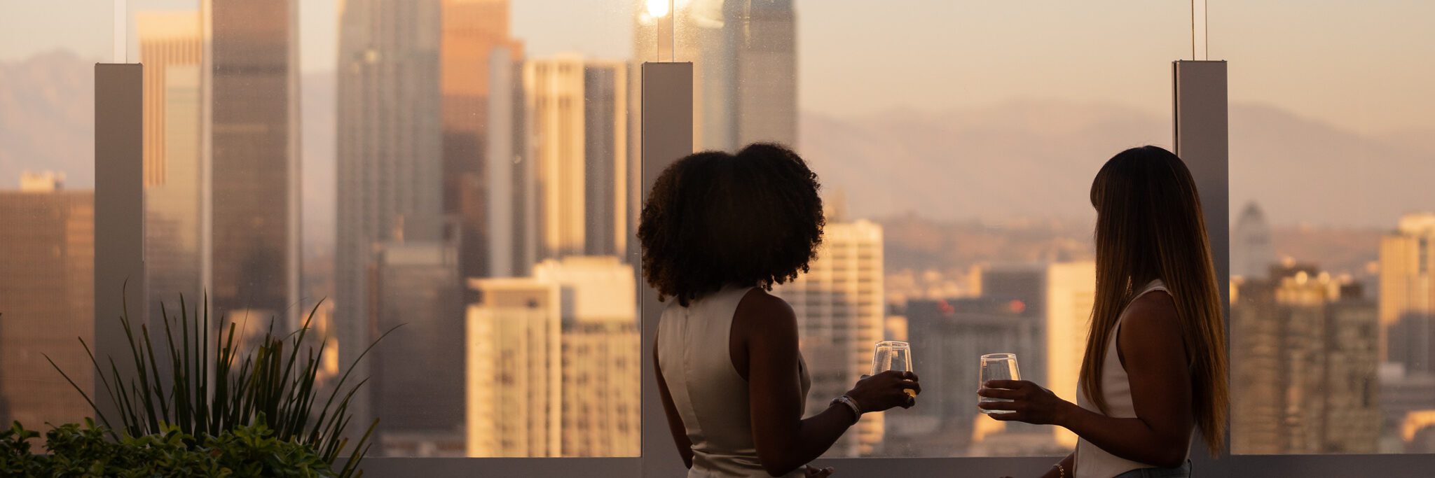 Two women dressed in light outfits stand on a rooftop, holding drinks and facing a city skyline at sunset, with tall buildings and mountains visible in the background.
