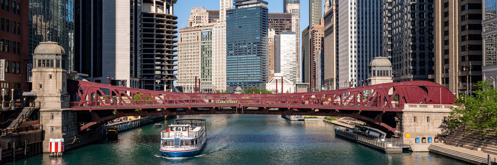 A boat travels on the Chicago River beneath a red steel bridge, surrounded by tall modern and historic skyscrapers on a clear sunny day.