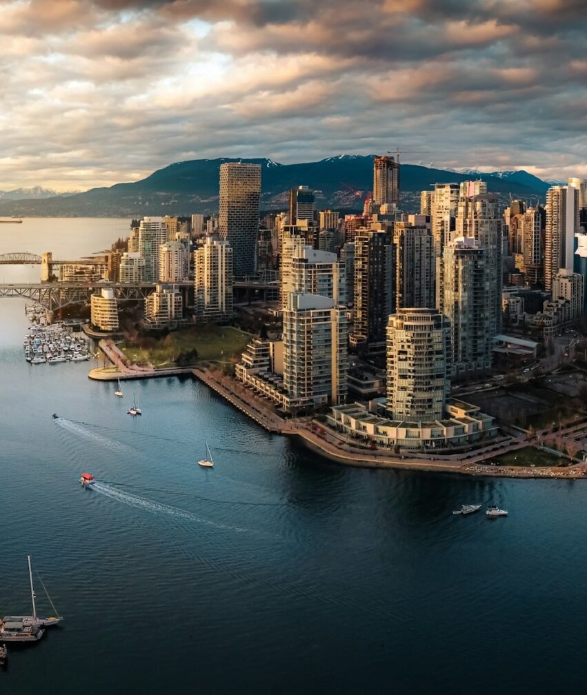 Aerial view of a modern city skyline by the water, with high-rise buildings, boats in the marina, and mountains in the background under a partly cloudy sky at sunset.