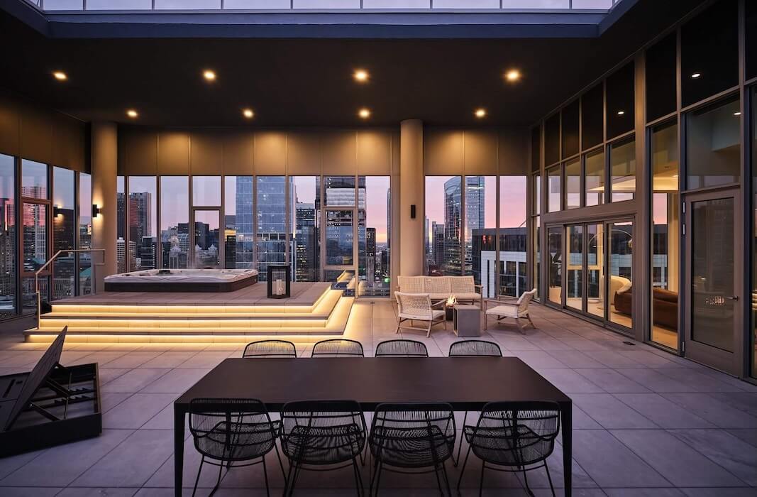 Modern rooftop terrace at sunset with city views, featuring a dining table, lounge chairs, a lit-up hot tub, and stylish outdoor seating, surrounded by tall glass windows and buildings.
