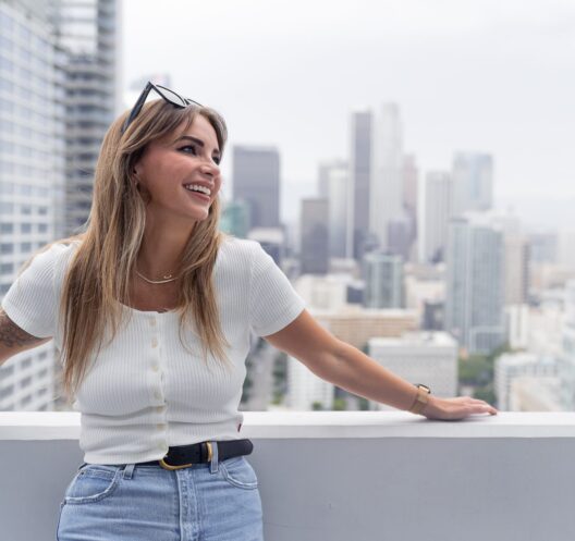 A woman with long hair, wearing a white top and blue jeans, smiles while leaning on a rooftop railing with a city skyline in the background—perfect for enjoying third night free hotel stays in Chicago, Los Angeles, and Seattle.